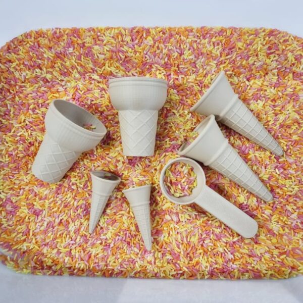 ijshoorn set Een kleurrijke ondergrond van roze, gele en oranje gekleurde rijst, waarop verschillende beige plastic ijshoornvormen en een schepje liggen – sensorisch speelmateriaal in ijsthema, geschikt voor fantasiespel en fijne motoriek